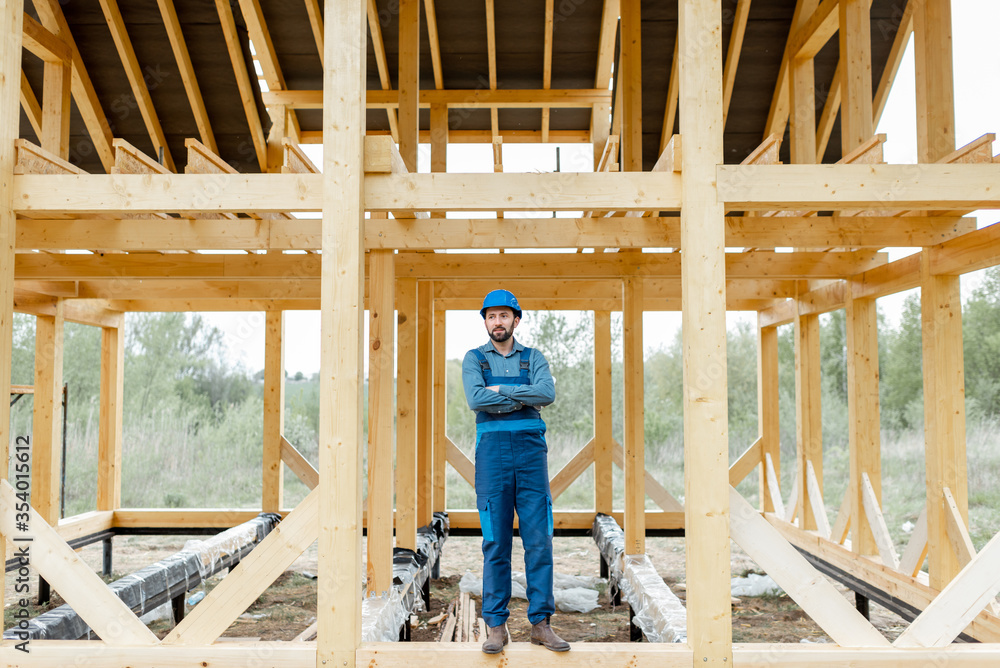 Builder in blue overalls and hard hat on the construction site, building wooden frame house Builder in blue overalls and hard hat on the construction site, building wooden frame house