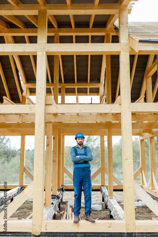 Builder in blue overalls and hard hat on the construction site, building wooden frame house Builder in blue overalls and hard hat on the construction site, building wooden frame house
