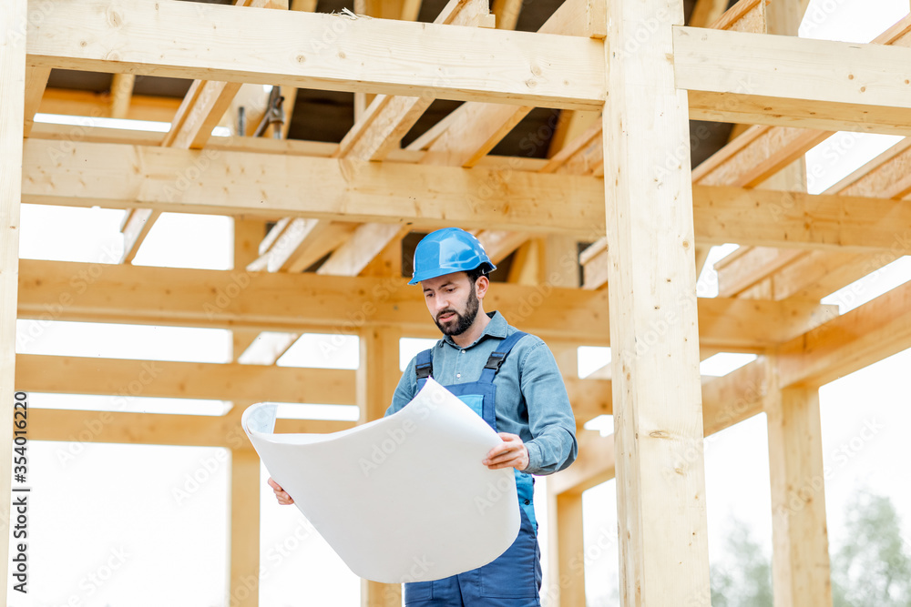 Builder in blue overalls and hard hat with blueprints on the construction site. Building wooden frame house concept