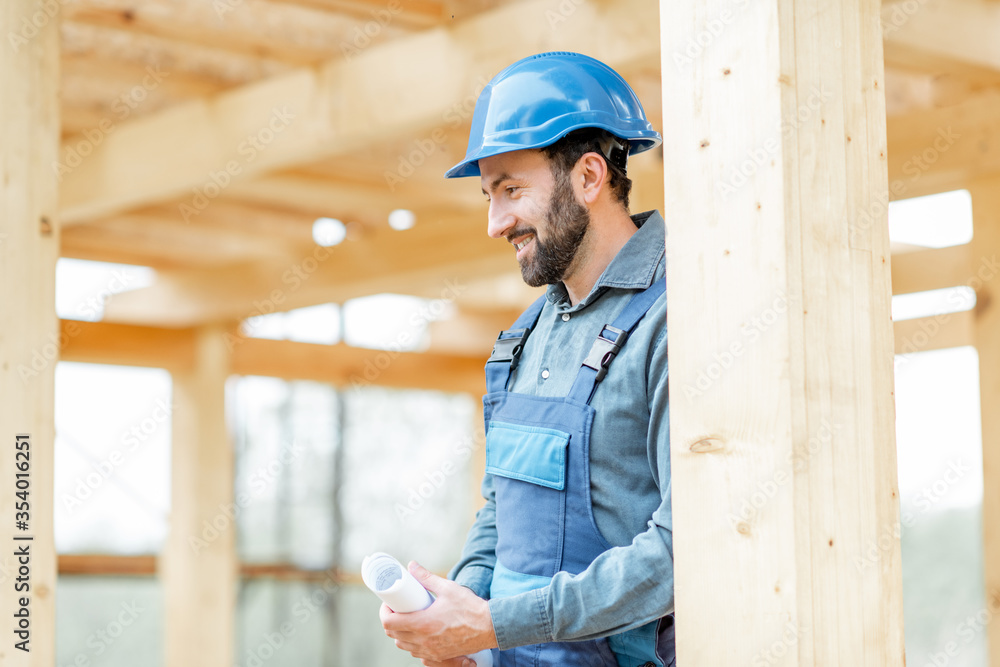 Portrait of a handsome and cofident builder in blue overalls and hard hat on the construction site. Building wooden frame house concept Portrait of a handsome and cofident builder in blue overalls and hard hat on the construction site. Building wooden frame house concept