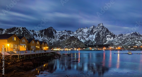 A panoramic evening shot of classic yellow rorbu reflecting the water under cloudy sky and snowy mountain in Ballstad, Lofoten island, Norway
