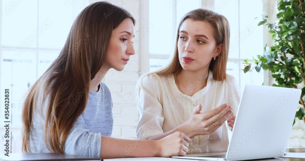 Two young concentrated female colleagues sharing ideas of project development, working together on laptop in modern office. Head shot focused business women discussing marketing strategy at workplace. Two young concentrated female colleagues sharing ideas of project development, working together on laptop in modern office. Head shot focused business women discussing marketing strategy at workplace.