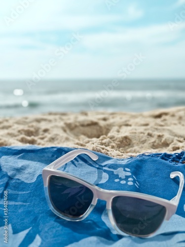 Sunglasses detail at beach. White sunglasses in foreground on blue towel, ocean and sand blurred in background. Summer holydays concept