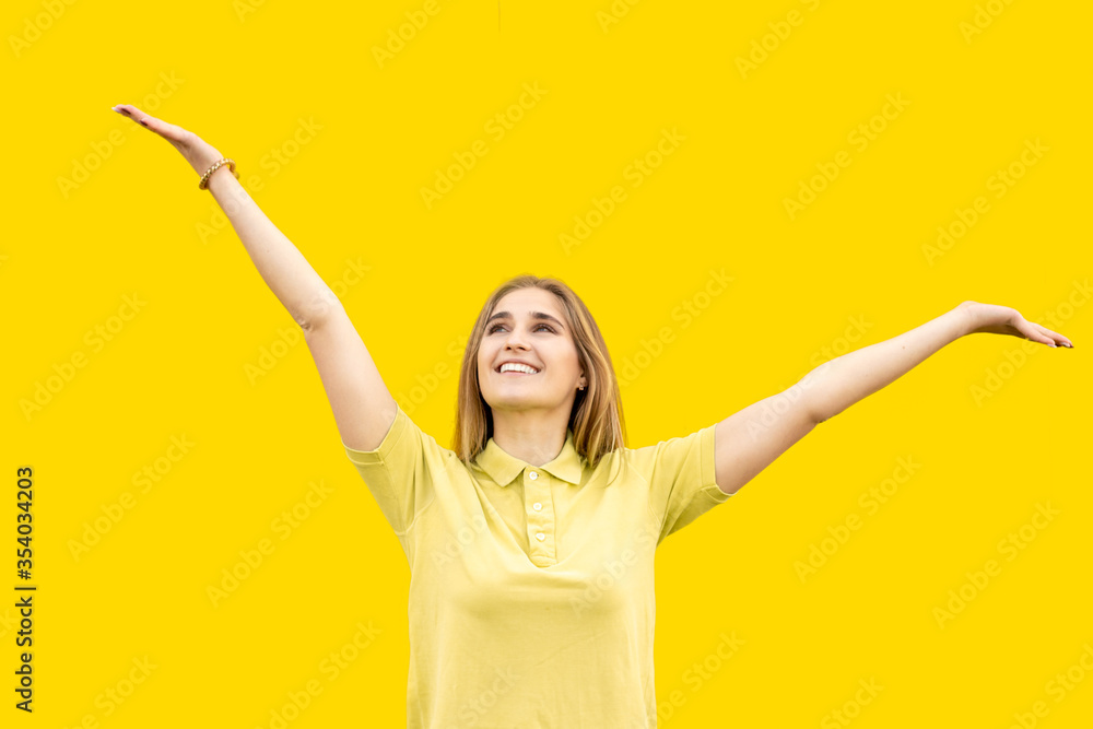 A young smiling girl is waving. Looks at something with a smile up. Dressed in a yellow T-shirt. Isolated on yellow background. A young smiling girl is waving. Looks at something with a smile up. Dressed in a yellow T-shirt. Isolated on yellow background.