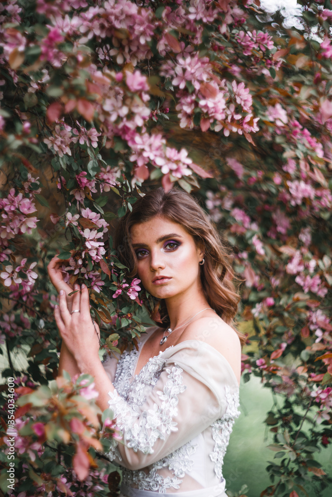A beautiful romantic girl in a white delicate dress stands near a Bush of pink flowers and touches a branch with flowers A beautiful romantic girl in a white delicate dress stands near a Bush of pink flowers and touches a branch with flowers