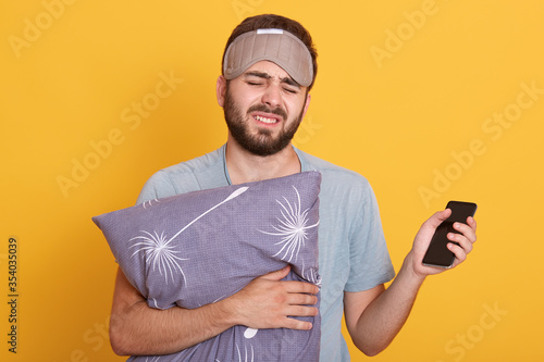 Photography Tired sleepy man with closed eyes wearing blindfold and t shirt, embracing gray pillow, unshaven guy with smart phone in hand, wants to go to bed, posing isolated over yellow background