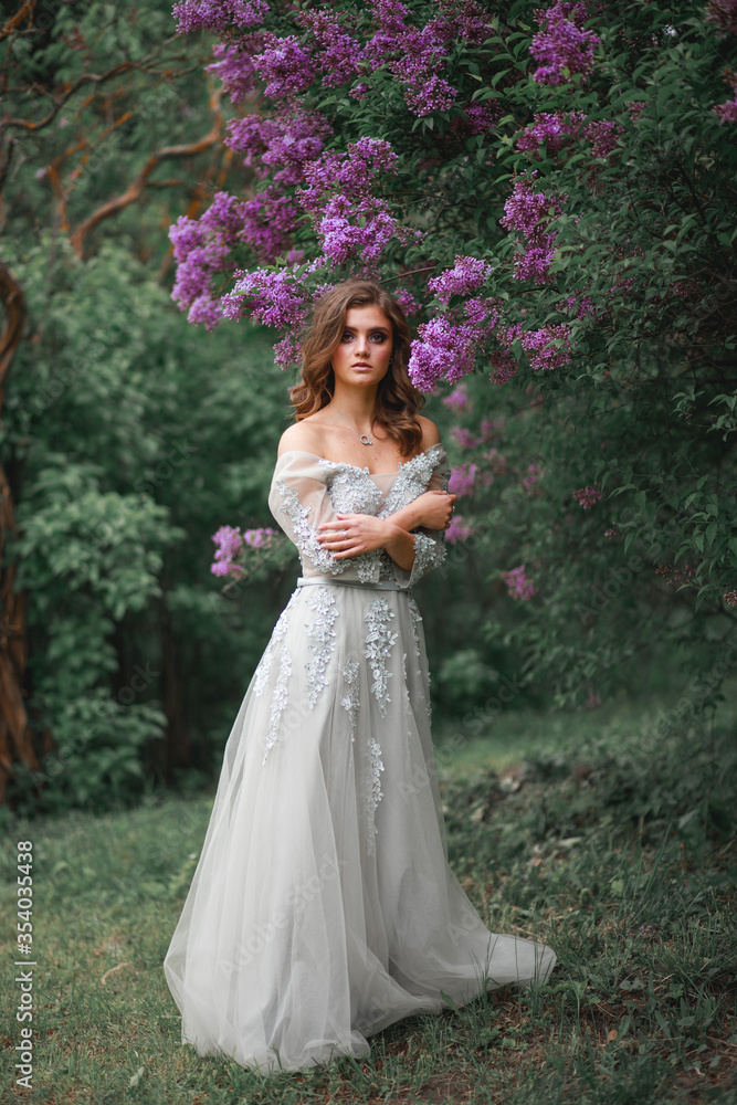 portrait of a girl in a white evening dress next to a lilac Bush
