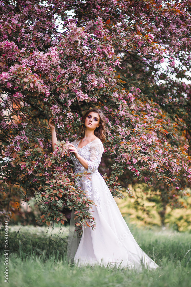 A beautiful romantic girl in a white delicate dress stands near a Bush of pink flowers and touches a branch with flowers