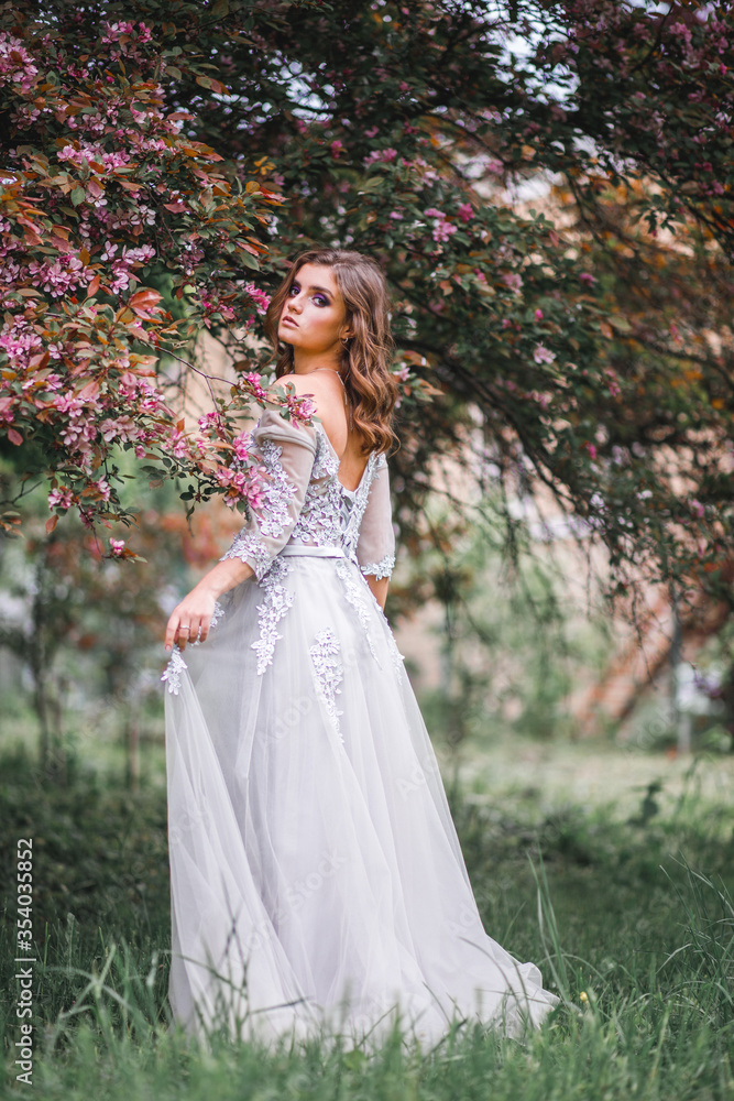 A beautiful romantic girl in a white delicate dress stands near a Bush of pink flowers and touches a branch with flowers
