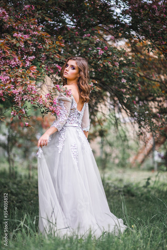 A beautiful romantic girl in a white delicate dress stands near a Bush of pink flowers and touches a branch with flowers © Aleksei Zakharov