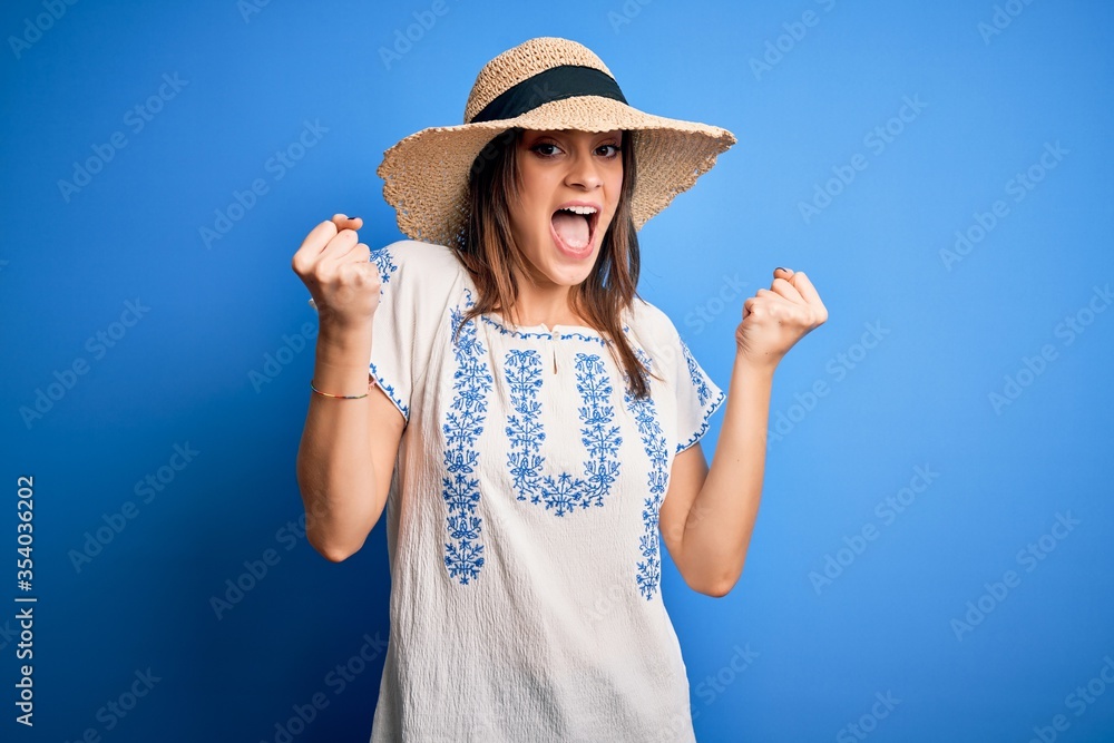 Young beautiful brunette woman wearing casual t-shirt and summer hat over blue background celebrating surprised and amazed for success with arms raised and open eyes. Winner concept.