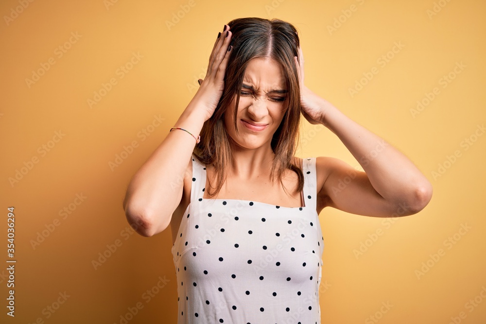 Young beautiful brunette woman wearing casual dress standing over yellow background suffering from headache desperate and stressed because pain and migraine. Hands on head.