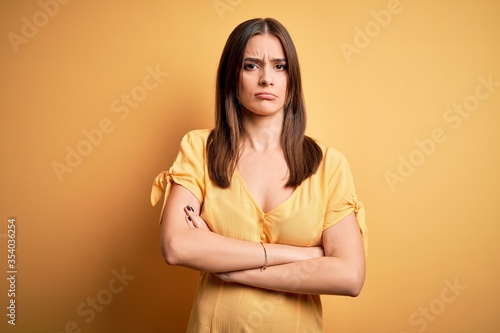 Young beautiful brunette woman wearing casual dress standing over yellow background skeptic and nervous, disapproving expression on face with crossed arms. Negative person.