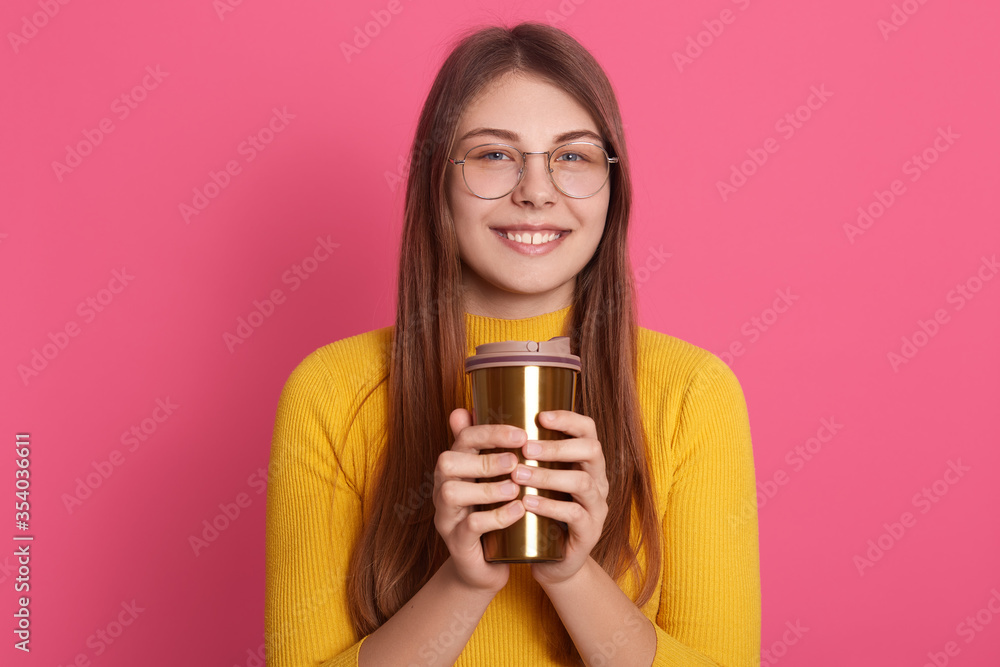 Portrait of relaxed young woman standing indoor and holding cup of coffee, looking directly at camera with charming smile, attractive female wearing yellow shirt and glasses, enjoys hot beverage. Portrait of relaxed young woman standing indoor and holding cup of coffee, looking directly at camera with charming smile, attractive female wearing yellow shirt and glasses, enjoys hot beverage.