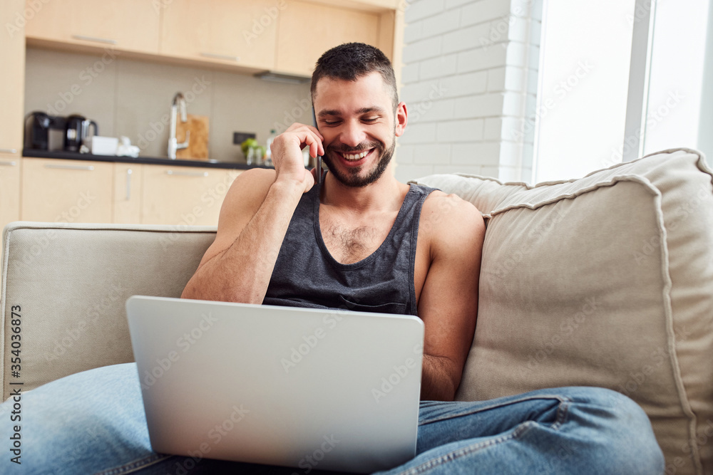 Fotografie Cheerful young man talking on cellphone and using laptop