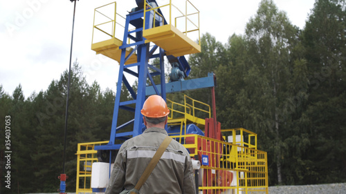 Oil field technical worker standing with his back to camera and in front of crude oil pump unit. Oilman in orange helmet overseeing site of crude oil production. Extraction of oil.