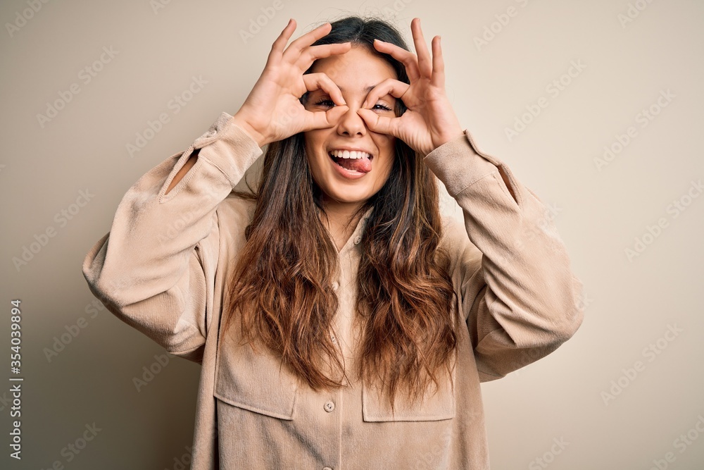 Young beautiful brunette woman wearing casual shirt standing over white background doing ok gesture like binoculars sticking tongue out, eyes looking through fingers. Crazy expression. Young beautiful brunette woman wearing casual shirt standing over white background doing ok gesture like binoculars sticking tongue out, eyes looking through fingers. Crazy expression.