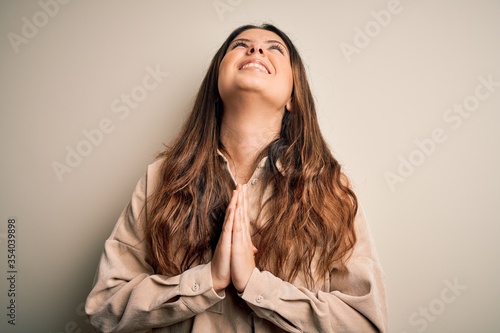 Young beautiful brunette woman wearing casual shirt standing over white background begging and praying with hands together with hope expression on face very emotional and worried. Begging.