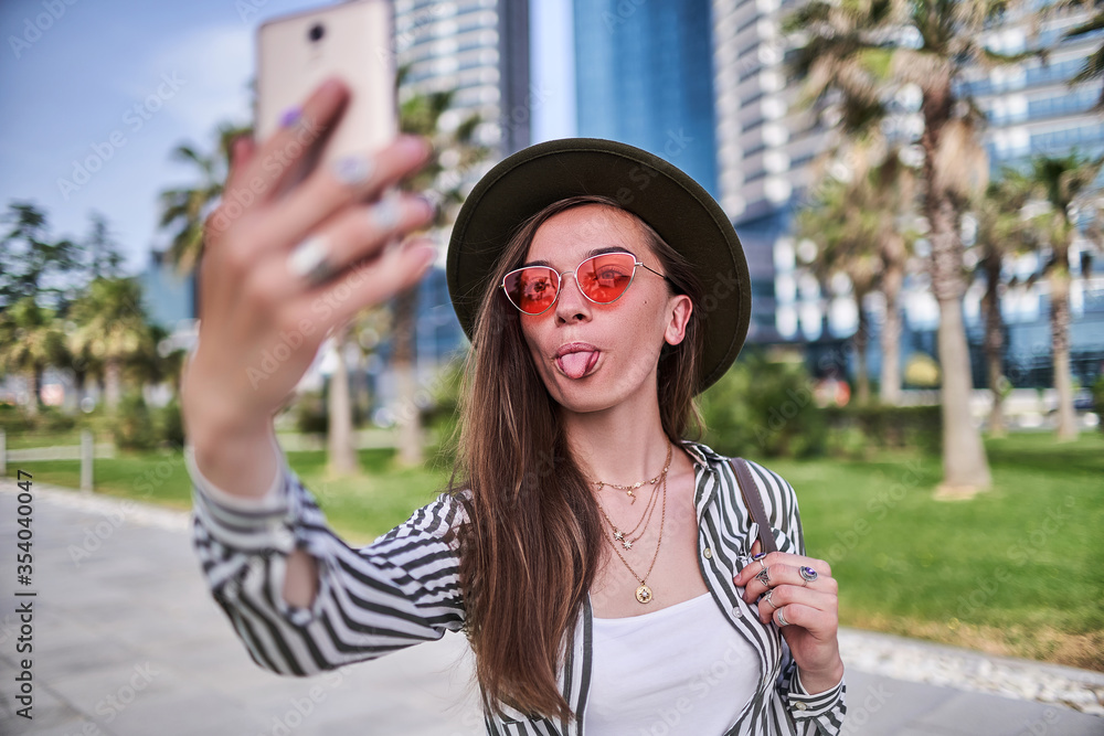 Gorgeous joyful funny modern carefree young hipster woman wearing felt hat and bright red glasses showing tongue during taking selfie photo portrait outdoors