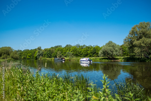 Idyllic scenic view of both a narrow boat and motor launch seen in an inland waterway in the UK. The wide expanse of the river is seen as well as the dense foliage and distant woodlands.