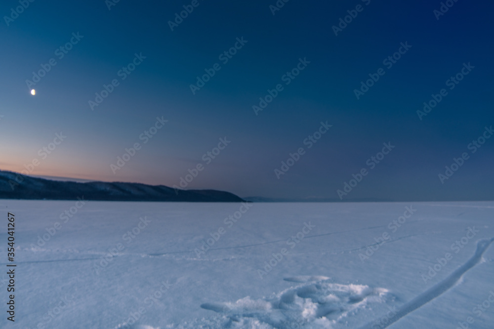 Snow-covered surface of Baikal Lake on the morning with track of bicycle and moon on the sky. Siberia, Russia Snow-covered surface of Baikal Lake on the morning with track of bicycle and moon on the sky. Siberia, Russia