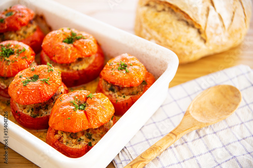 Mediterranean stuffed tomatoes with meat, bread crumbs, and herbs in a white oven dish, aside a kitchen towel, a bread loaf and a wooden serving spoon on an oak wood table.