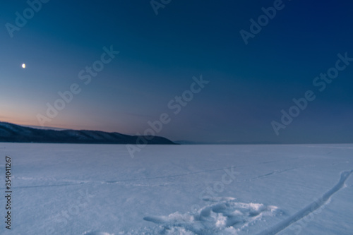 Snow-covered surface of Baikal Lake on the morning with track of bicycle and moon on the sky. Siberia, Russia © evdokimari