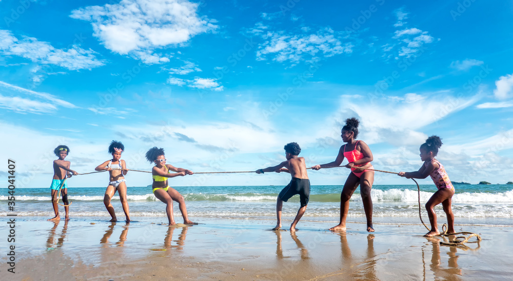 A group of children pulling rope and competing on the beach against blue sky.