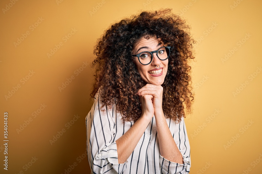 Young beautiful woman with curly hair and piercing wearing striped shirt and glasses laughing nervous and excited with hands on chin looking to the side Young beautiful woman with curly hair and piercing wearing striped shirt and glasses laughing nervous and excited with hands on chin looking to the side