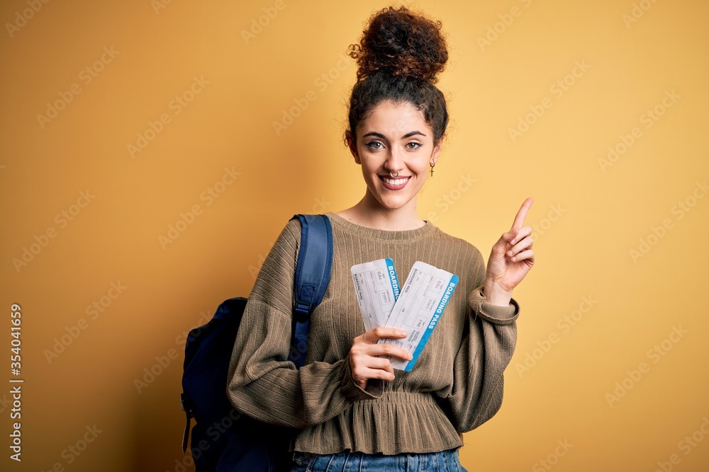 Beautiful tourist woman with curly hair and piercing wearing backpack holding boarding pass surprised with an idea or question pointing finger with happy face, number one Beautiful tourist woman with curly hair and piercing wearing backpack holding boarding pass surprised with an idea or question pointing finger with happy face, number one