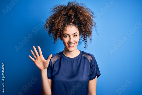 Young beautiful woman with curly hair and piercing wearing casual blue t-shirt showing and pointing up with fingers number five while smiling confident and happy.