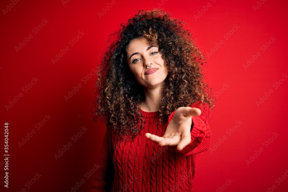 Young beautiful woman with curly hair and piercing wearing casual red sweater smiling cheerful offering palm hand giving assistance and acceptance. Young beautiful woman with curly hair and piercing wearing casual red sweater smiling cheerful offering palm hand giving assistance and acceptance.