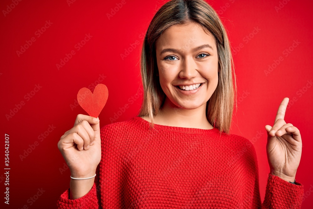 Young beautiful woman holding heart card standing over isolated red background surprised with an idea or question pointing finger with happy face, number one Young beautiful woman holding heart card standing over isolated red background surprised with an idea or question pointing finger with happy face, number one