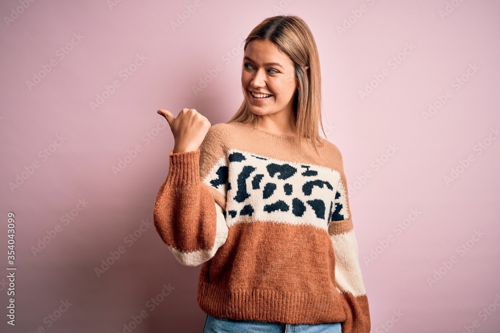 Young beautiful blonde woman wearing fashion animal print sweater over pink background smiling with happy face looking and pointing to the side with thumb up. Young beautiful blonde woman wearing fashion animal print sweater over pink background smiling with happy face looking and pointing to the side with thumb up.