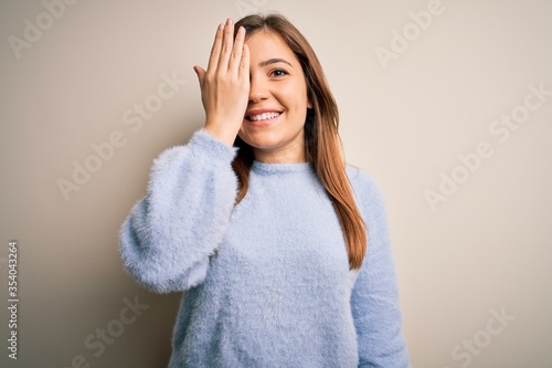 Beautiful young woman wearing casual winter sweater standing over isolated background covering one eye with hand, confident smile on face and surprise emotion.
