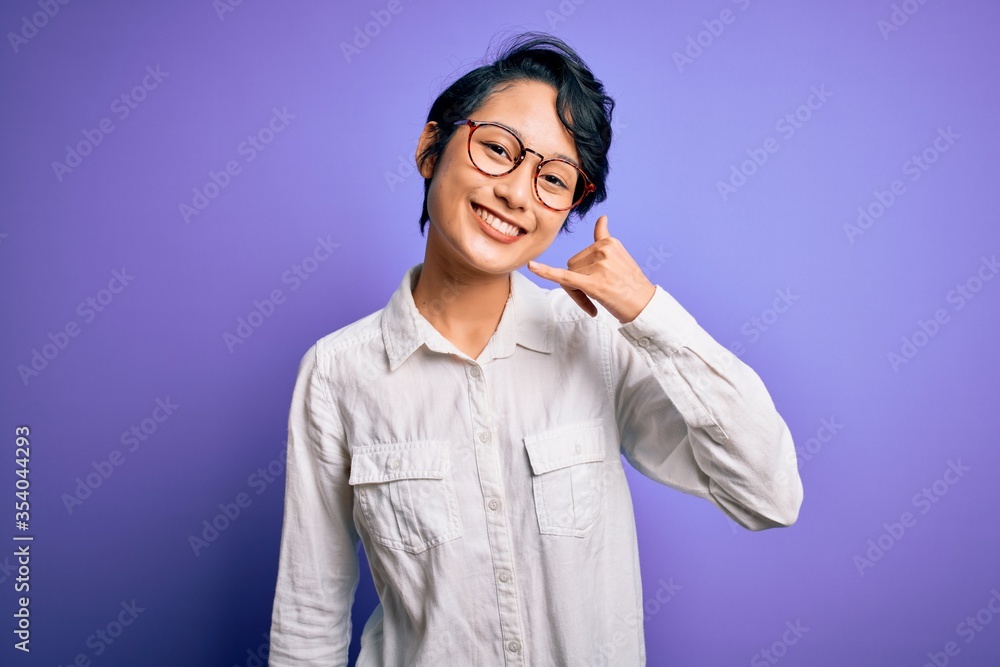 Young beautiful asian girl wearing casual shirt and glasses standing over purple background smiling doing phone gesture with hand and fingers like talking on the telephone. Communicating concepts. Young beautiful asian girl wearing casual shirt and glasses standing over purple background smiling doing phone gesture with hand and fingers like talking on the telephone. Communicating concepts.