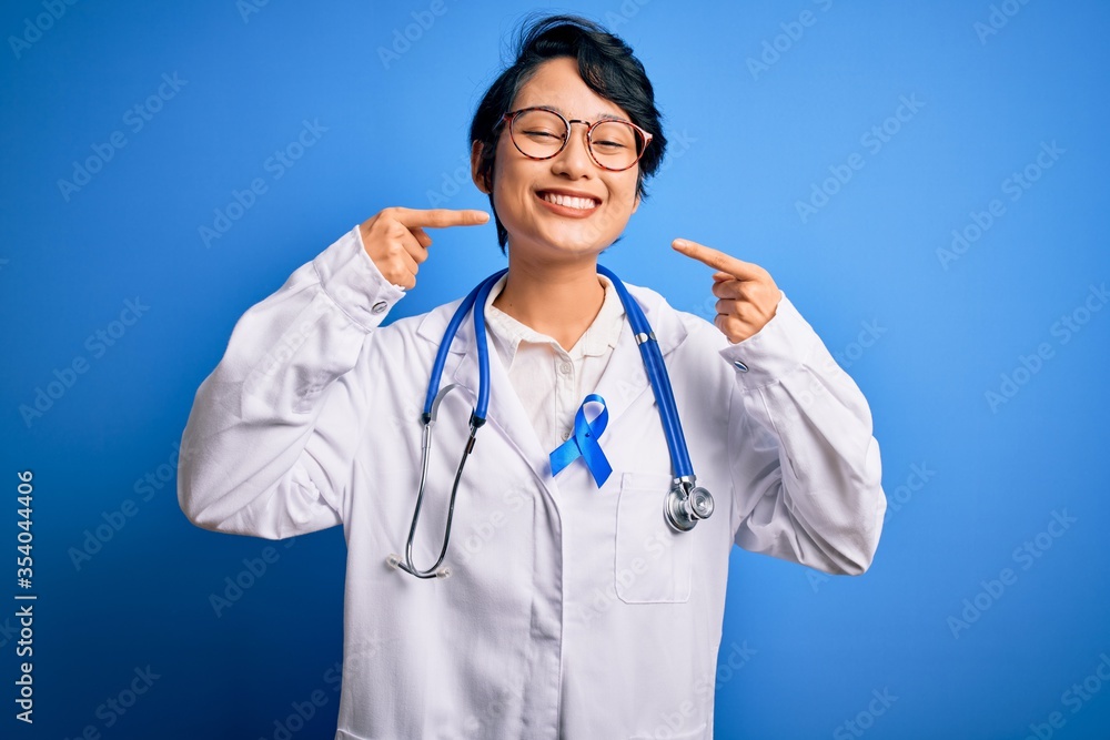 Young beautiful asian doctor girl wearing stethoscope and coat with blue cancer ribbon smiling cheerful showing and pointing with fingers teeth and mouth. Dental health concept. Young beautiful asian doctor girl wearing stethoscope and coat with blue cancer ribbon smiling cheerful showing and pointing with fingers teeth and mouth. Dental health concept.