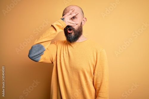 Handsome bald man with beard wearing casual sweater standing over yellow background peeking in shock covering face and eyes with hand, looking through fingers with embarrassed expression.