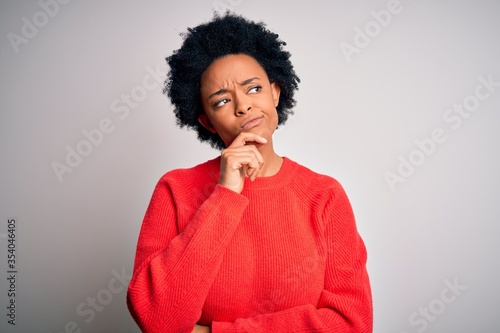 Young beautiful African American afro woman with curly hair wearing red casual sweater with hand on chin thinking about question  pensive expression. Smiling with thoughtful face. Doubt concept.