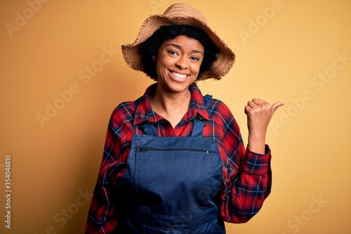 Young African American afro farmer woman with curly hair wearing apron and hat smiling with happy face looking and pointing to the side with thumb up.