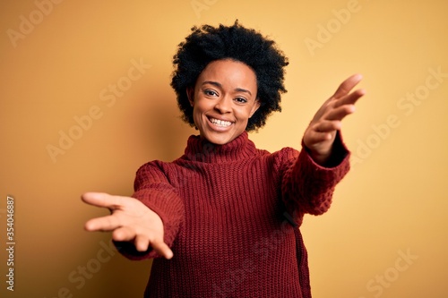 Young beautiful African American afro woman with curly hair wearing casual turtleneck sweater smiling cheerful offering hands giving assistance and acceptance.