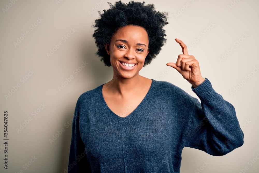Young beautiful African American afro woman with curly hair wearing casual sweater smiling and confident gesturing with hand doing small size sign with fingers looking and the camera. Measure concept. Young beautiful African American afro woman with curly hair wearing casual sweater smiling and confident gesturing with hand doing small size sign with fingers looking and the camera. Measure concept.