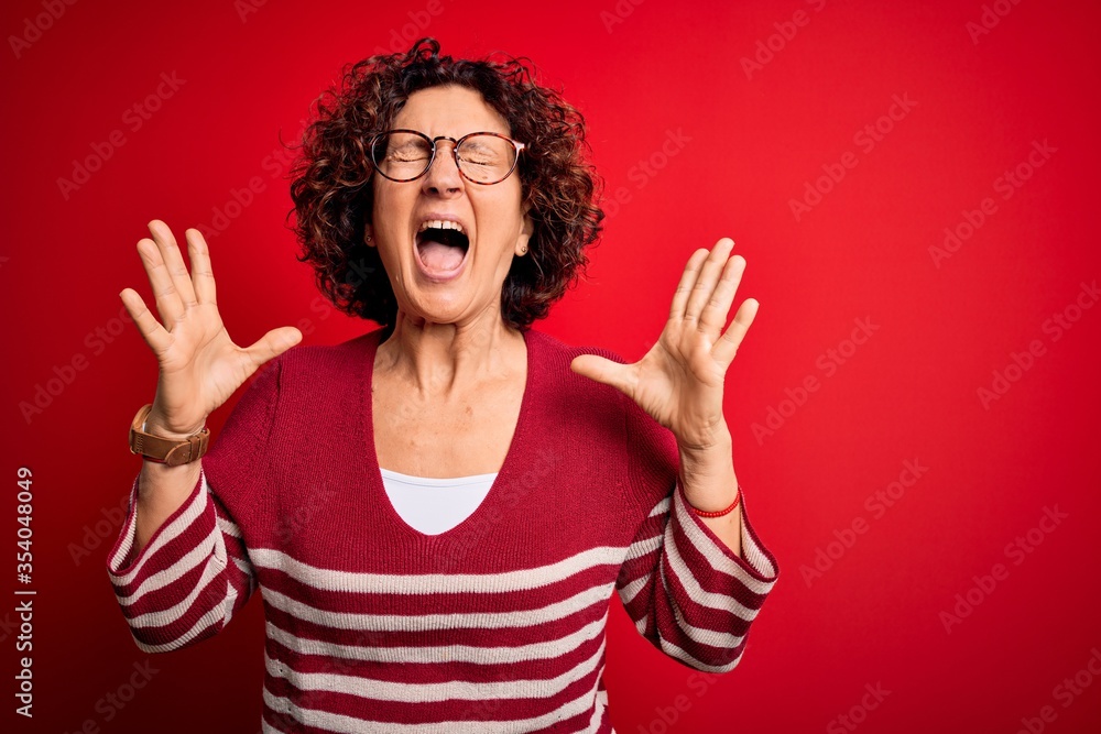 Middle age beautiful curly hair woman wearing casual striped sweater over red background celebrating mad and crazy for success with arms raised and closed eyes screaming excited. Winner concept Middle age beautiful curly hair woman wearing casual striped sweater over red background celebrating mad and crazy for success with arms raised and closed eyes screaming excited. Winner concept