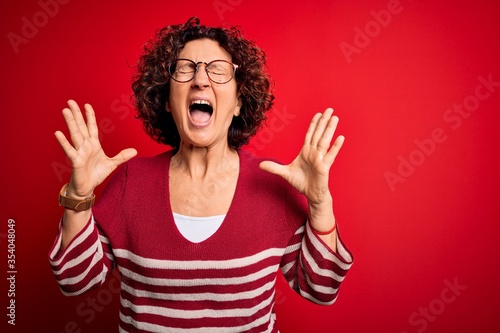 Middle age beautiful curly hair woman wearing casual striped sweater over red background celebrating mad and crazy for success with arms raised and closed eyes screaming excited. Winner concept