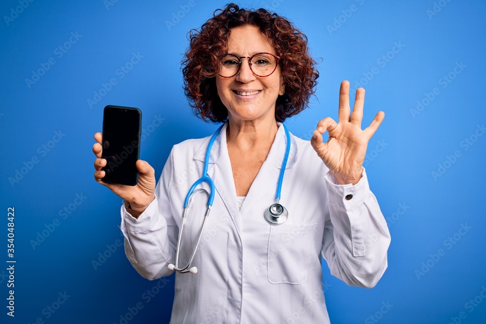 Middle age doctor woman wearing coat and stethoscope holding smartphone showing screen doing ok sign with fingers, excellent symbol Middle age doctor woman wearing coat and stethoscope holding smartphone showing screen doing ok sign with fingers, excellent symbol