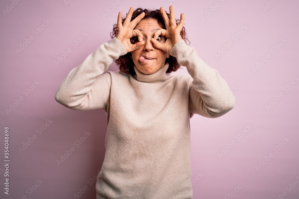 Middle age beautiful curly hair woman wearing casual turtleneck sweater over pink background doing ok gesture like binoculars sticking tongue out, eyes looking through fingers. Crazy expression. Middle age beautiful curly hair woman wearing casual turtleneck sweater over pink background doing ok gesture like binoculars sticking tongue out, eyes looking through fingers. Crazy expression.