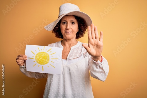 Middle age curly woman on vacation holding bunner with sun image over yellow background with open hand doing stop sign with serious and confident expression, defense gesture