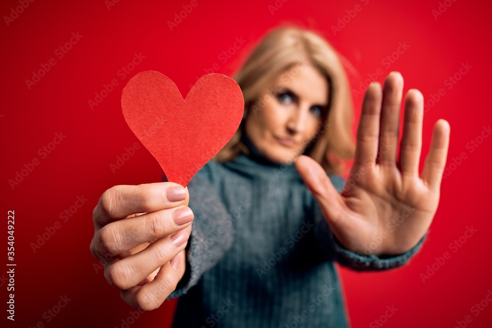 Middle age beautiful blonde romantic woman holding red paper heart celebrating valentine day with open hand doing stop sign with serious and confident expression, defense gesture Middle age beautiful blonde romantic woman holding red paper heart celebrating valentine day with open hand doing stop sign with serious and confident expression, defense gesture