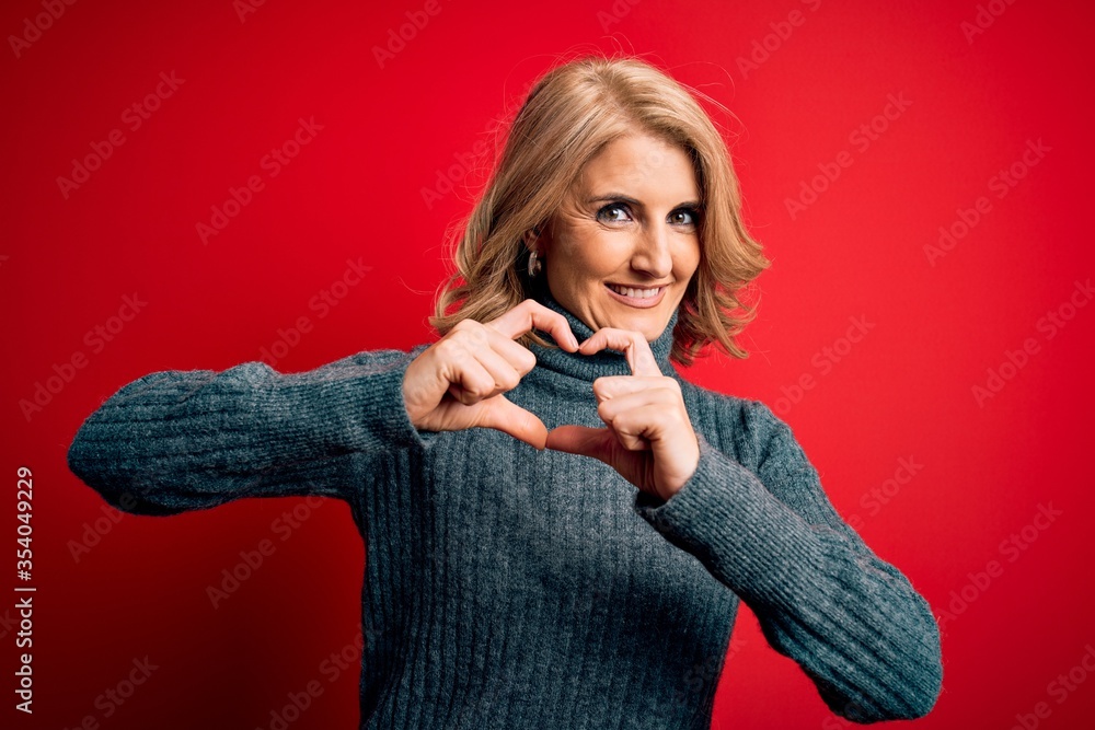 Naklejka premium Middle age beautiful blonde woman wearing casual turtleneck sweater over red background smiling in love showing heart symbol and shape with hands. Romantic concept.