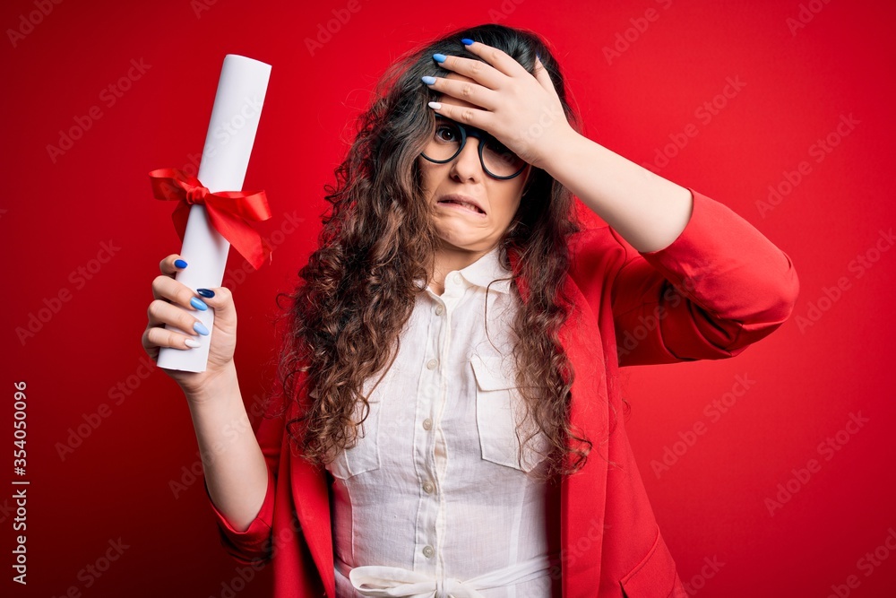 Young beautiful woman with curly hair holding university diploma degree over red background stressed with hand on head, shocked with shame and surprise face, angry and frustrated. Fear and upset Young beautiful woman with curly hair holding university diploma degree over red background stressed with hand on head, shocked with shame and surprise face, angry and frustrated. Fear and upset